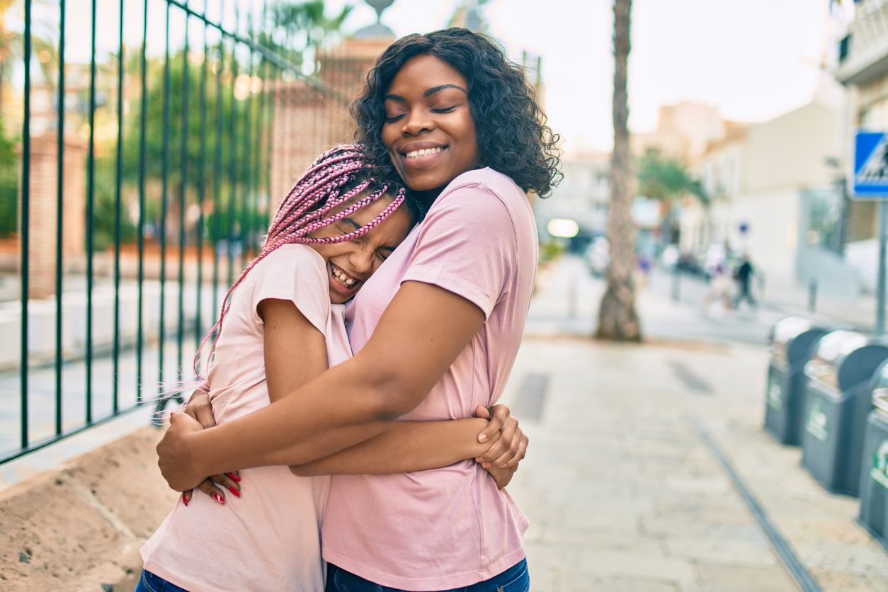 Mom hugs her daughter while standing on a sidewalk. Both are wearing pink shirts.