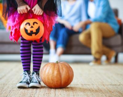 Child in costume holding a pumpkin basket with parents sitting on couch in background